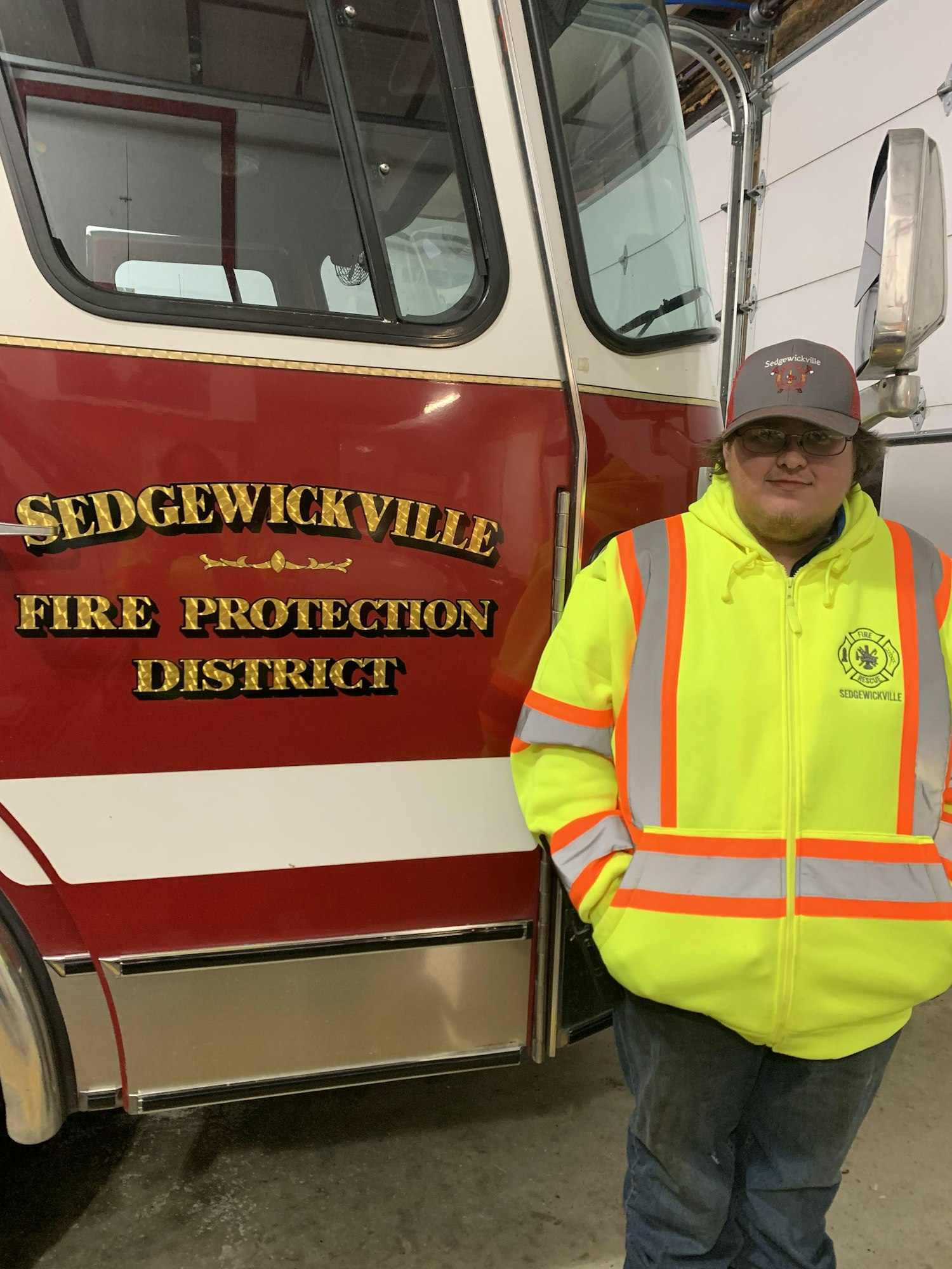 Person in a yellow reflective jacket stands beside a Sedgwickville Fire Protection District fire truck.