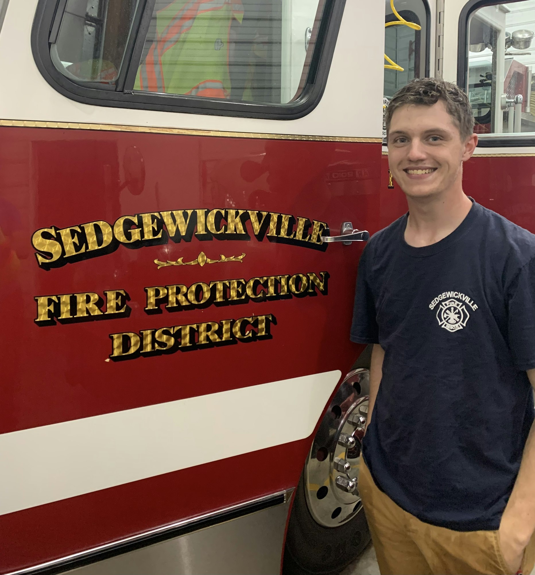 A smiling person in front of a fire truck with "Sedgewickville Fire Protection District" on the side.