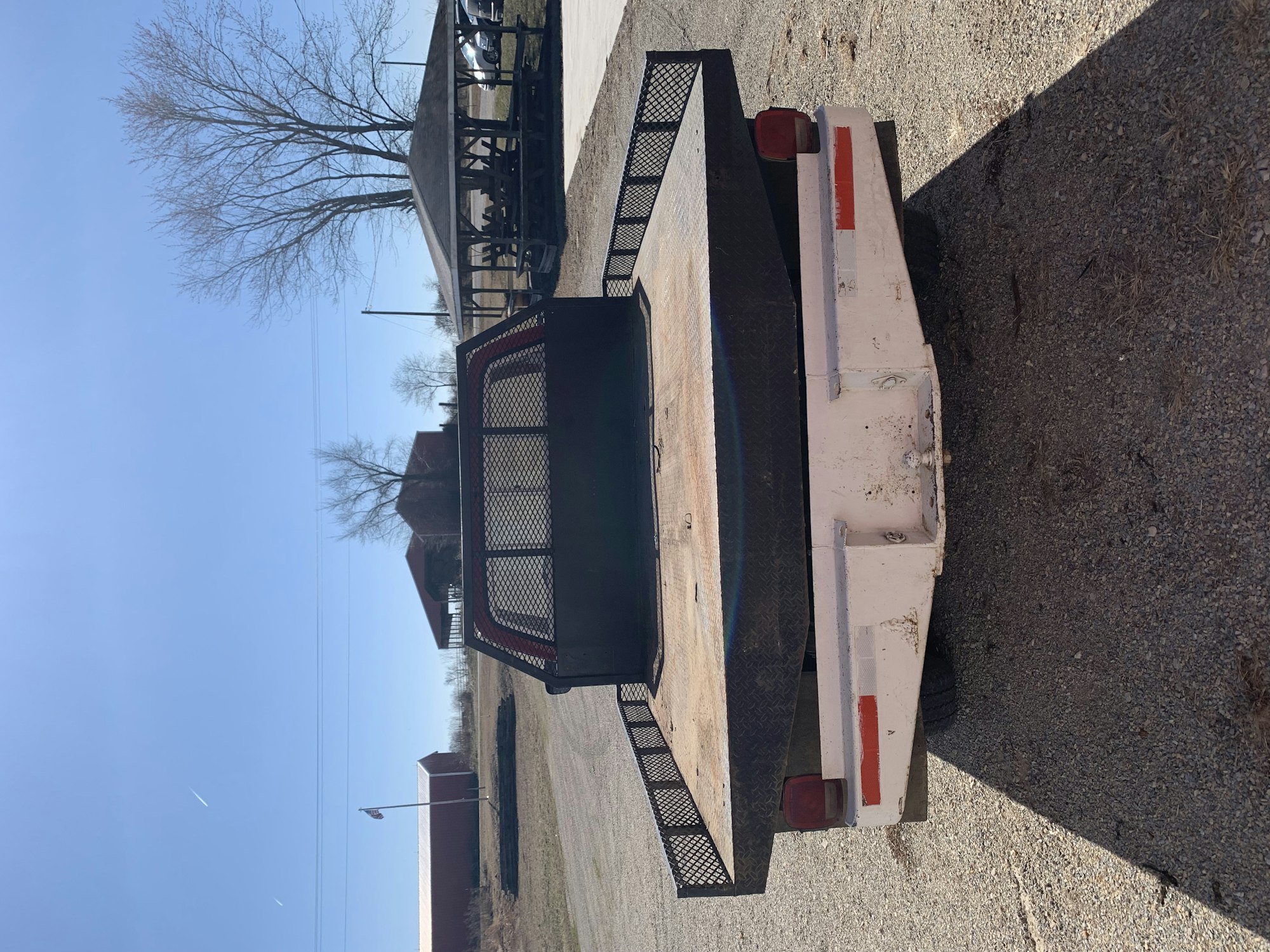A utility trailer parked on a gravel surface, image rotated counterclockwise, clear skies, and bare trees in the background.