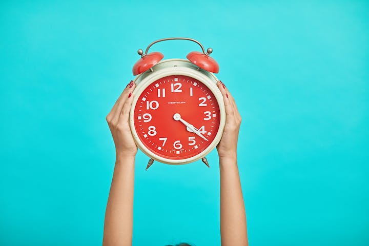 Two hands holding a red alarm clock up in front of a teal background.