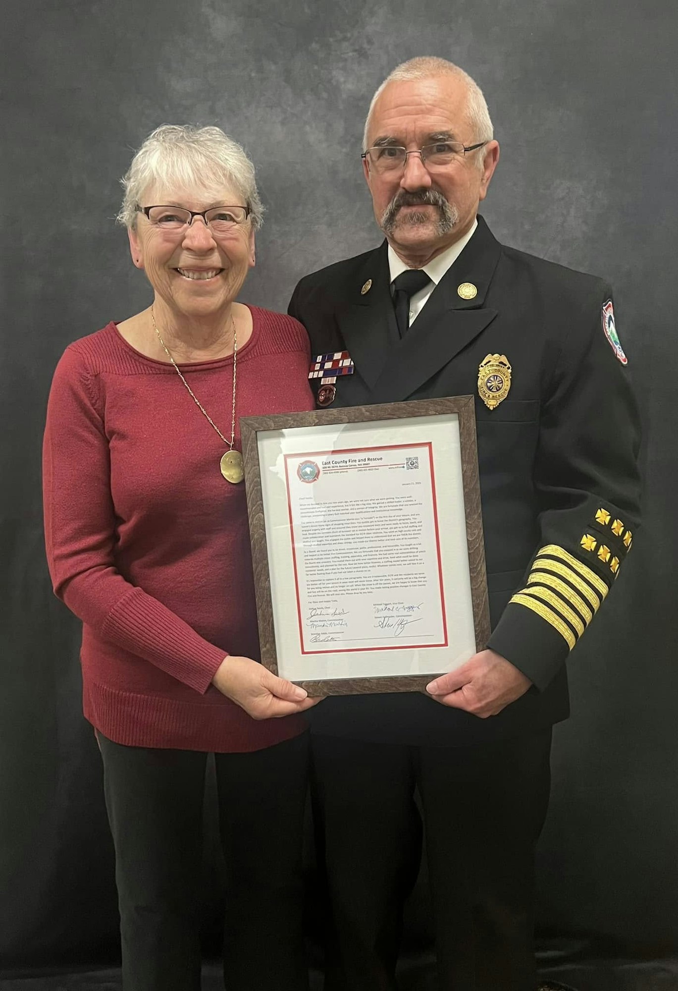 Two people standing together, one in a uniform holding a framed certificate, smiling against a plain background.
