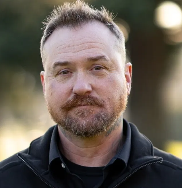 A man with a beard and short hair is looking directly at the camera, wearing a black jacket outdoors.