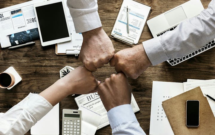Four hands are fist-bumping over a cluttered desk with papers, a calculator, and electronic devices, symbolizing teamwork.