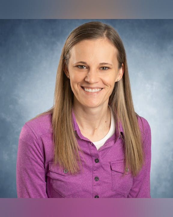 A person with long hair wearing a purple shirt, smiling against a blue background.