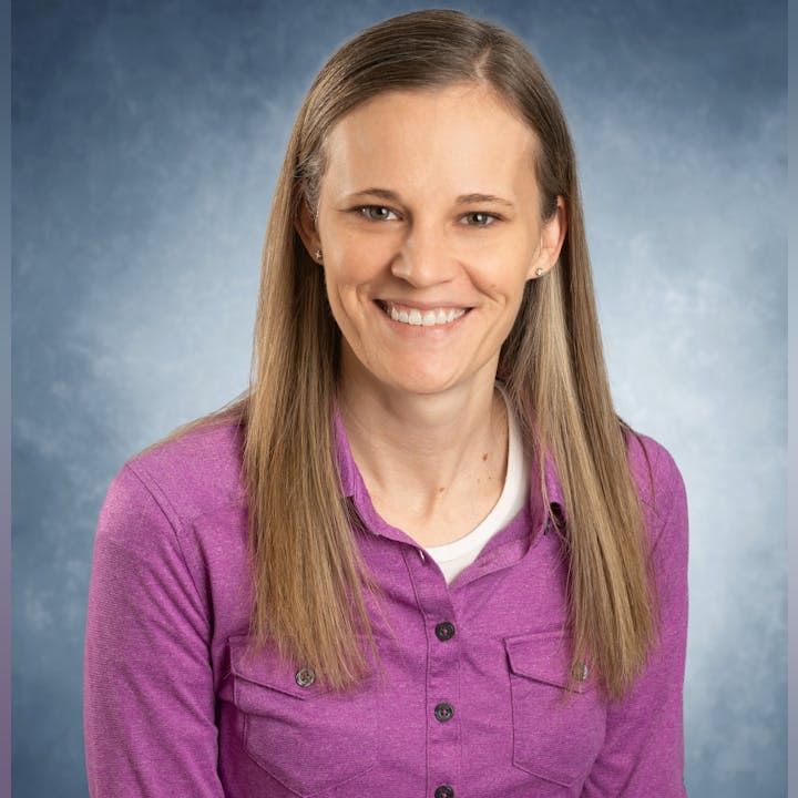 A person with long hair wearing a purple shirt, smiling against a blue background.
