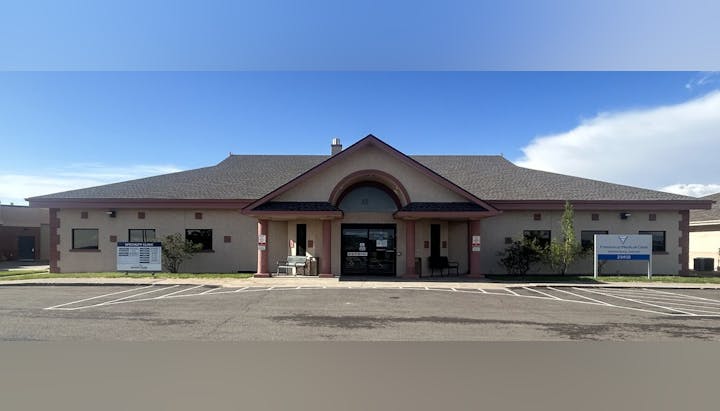 A single-story medical clinic building with parking spaces, signs, and clear sky.