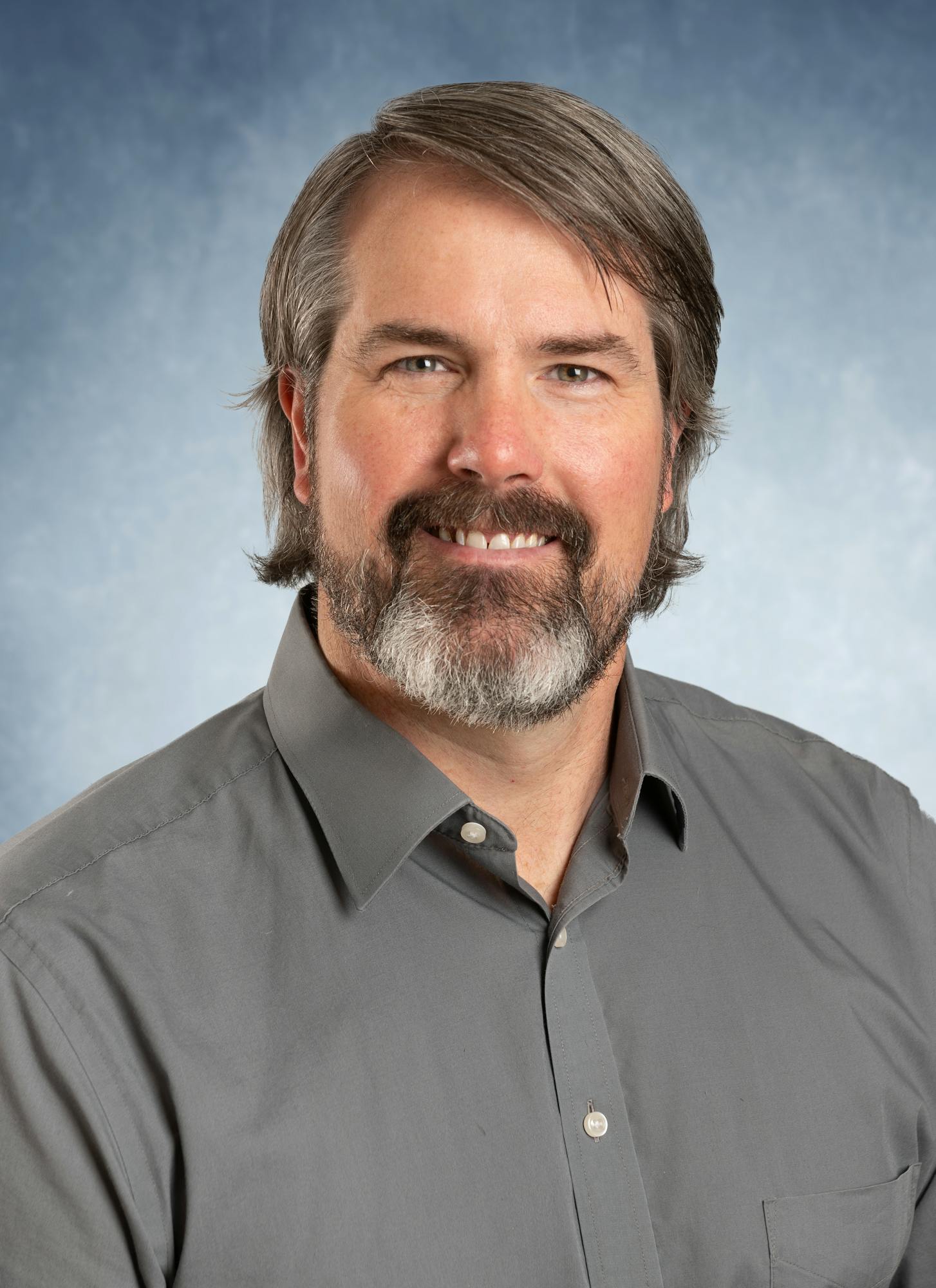 A man with a beard and gray shirt against a blue background.