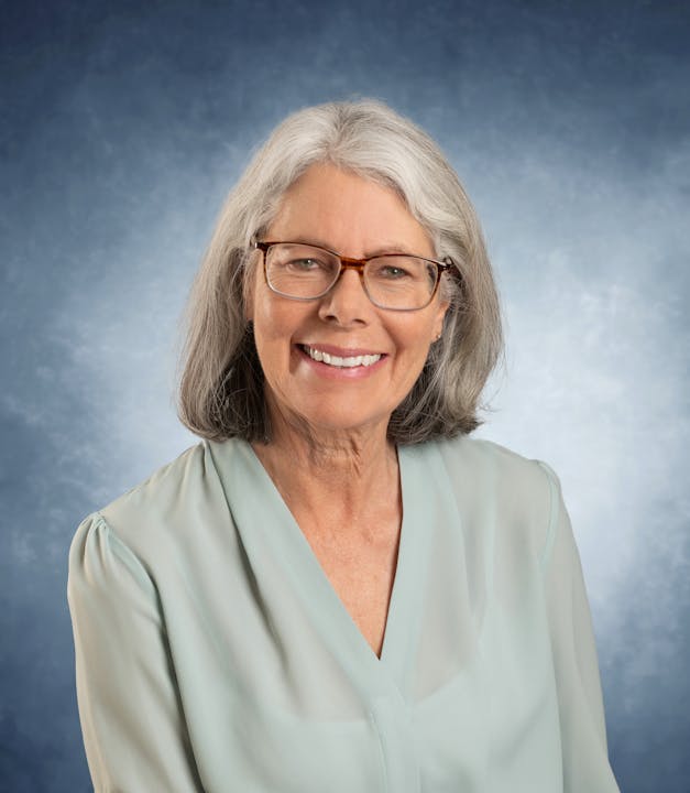 A woman with glasses and gray hair smiling, wearing a light blue blouse against a blue background.
