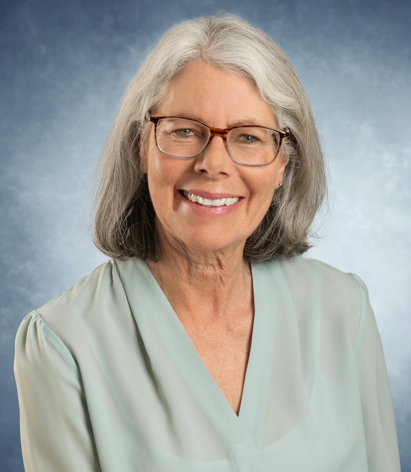 A woman with glasses and gray hair smiling, wearing a light blue blouse against a blue background.