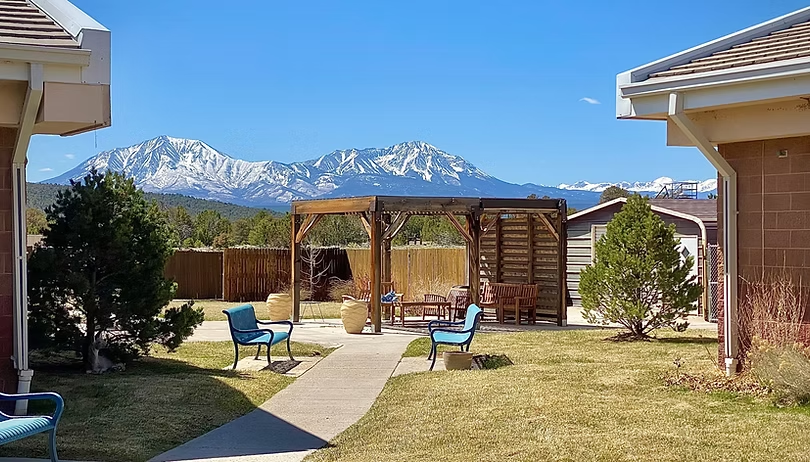 Snowy mountains, clear sky, a wooden pergola, seating area, and trees in a grassy courtyard between buildings.