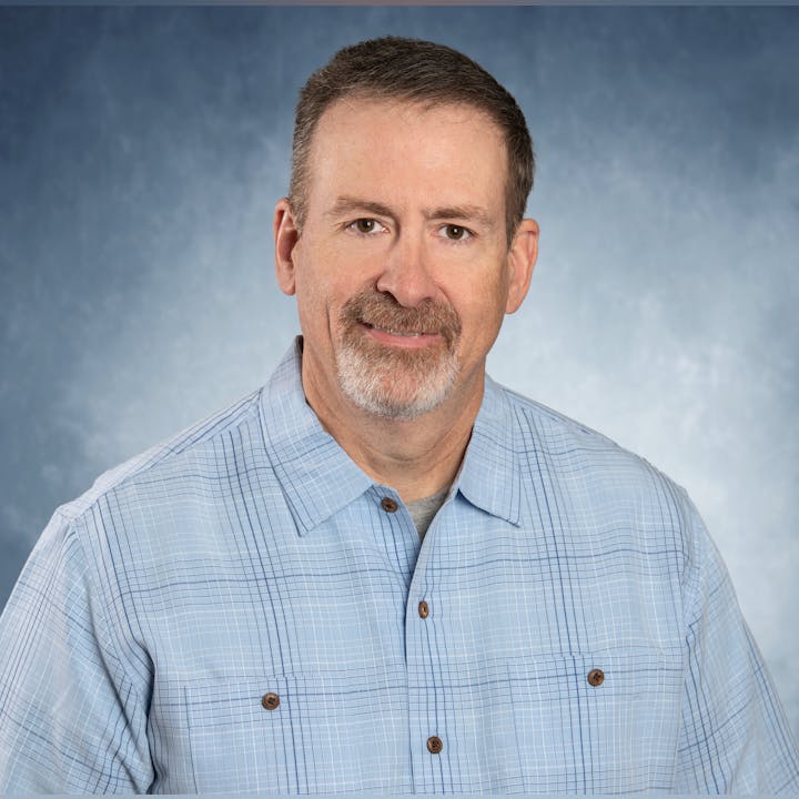 A man with short hair and a beard, wearing a light blue checkered shirt, smiling against a gray-blue background.