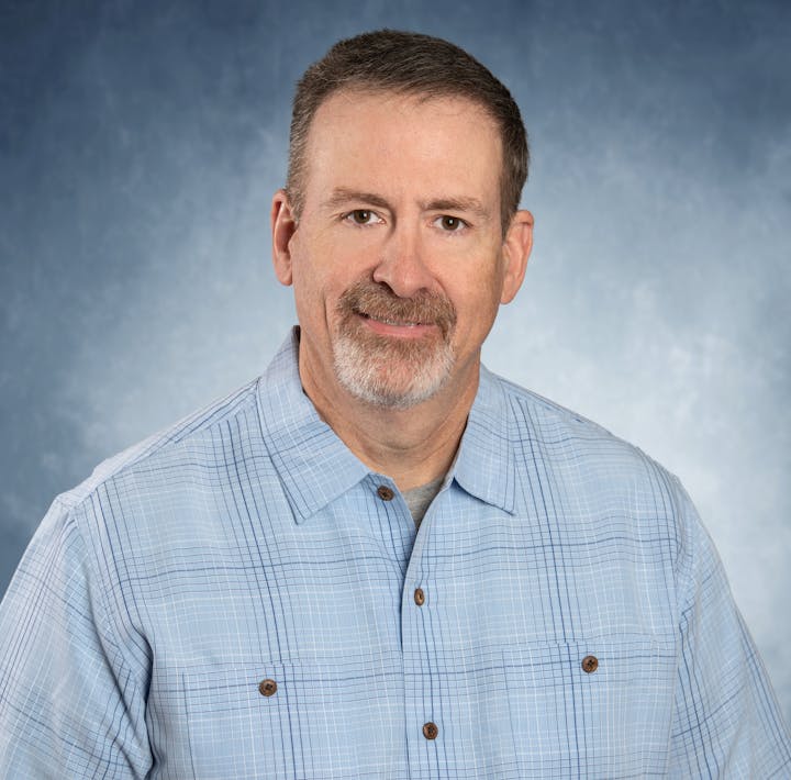 A man with short hair and a beard, wearing a light blue checkered shirt, smiling against a gray-blue background.
