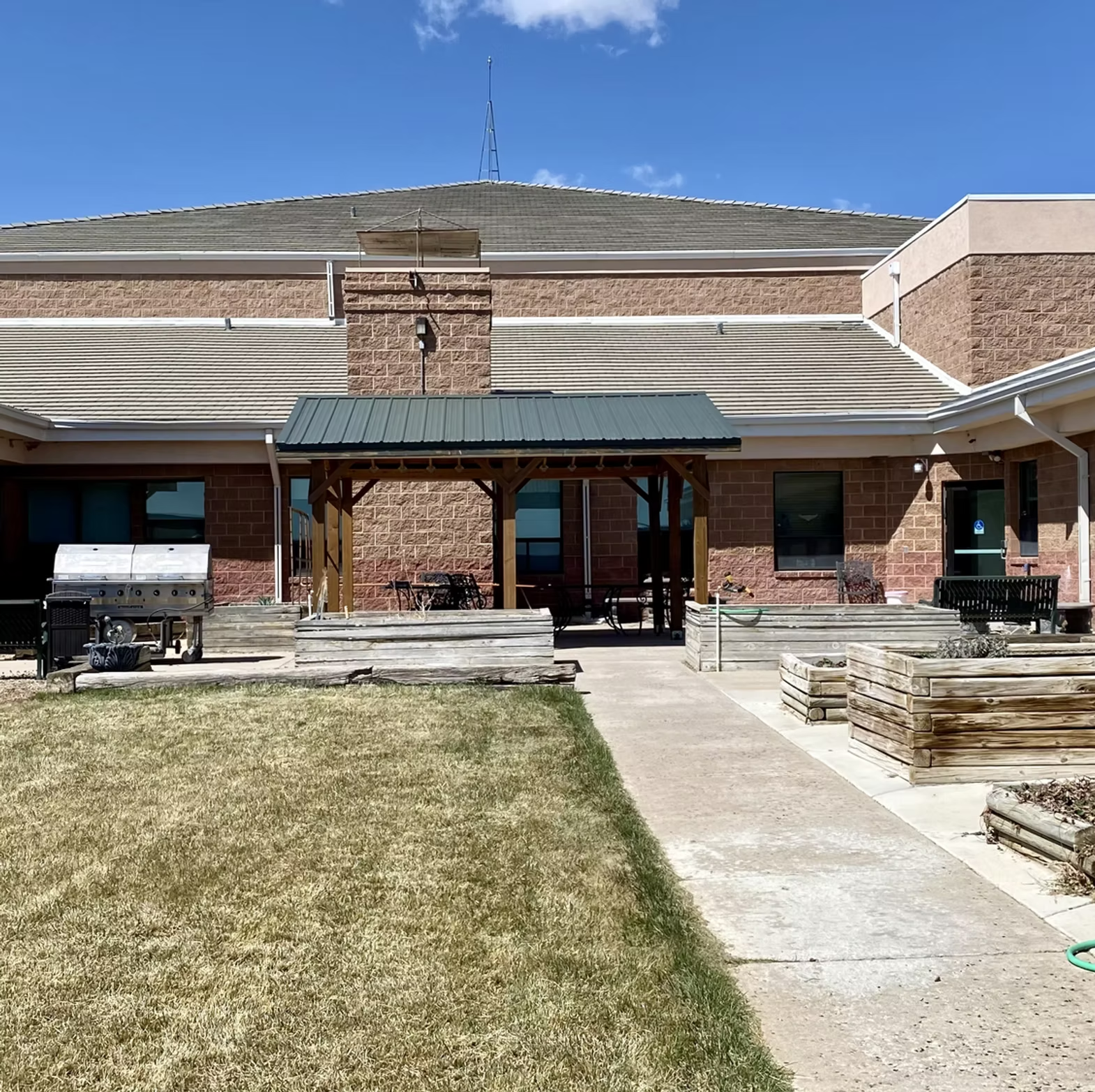 A brick building with a green-roofed canopy, a grassy yard, plant boxes, and a grill in a sunny, clear sky setting.