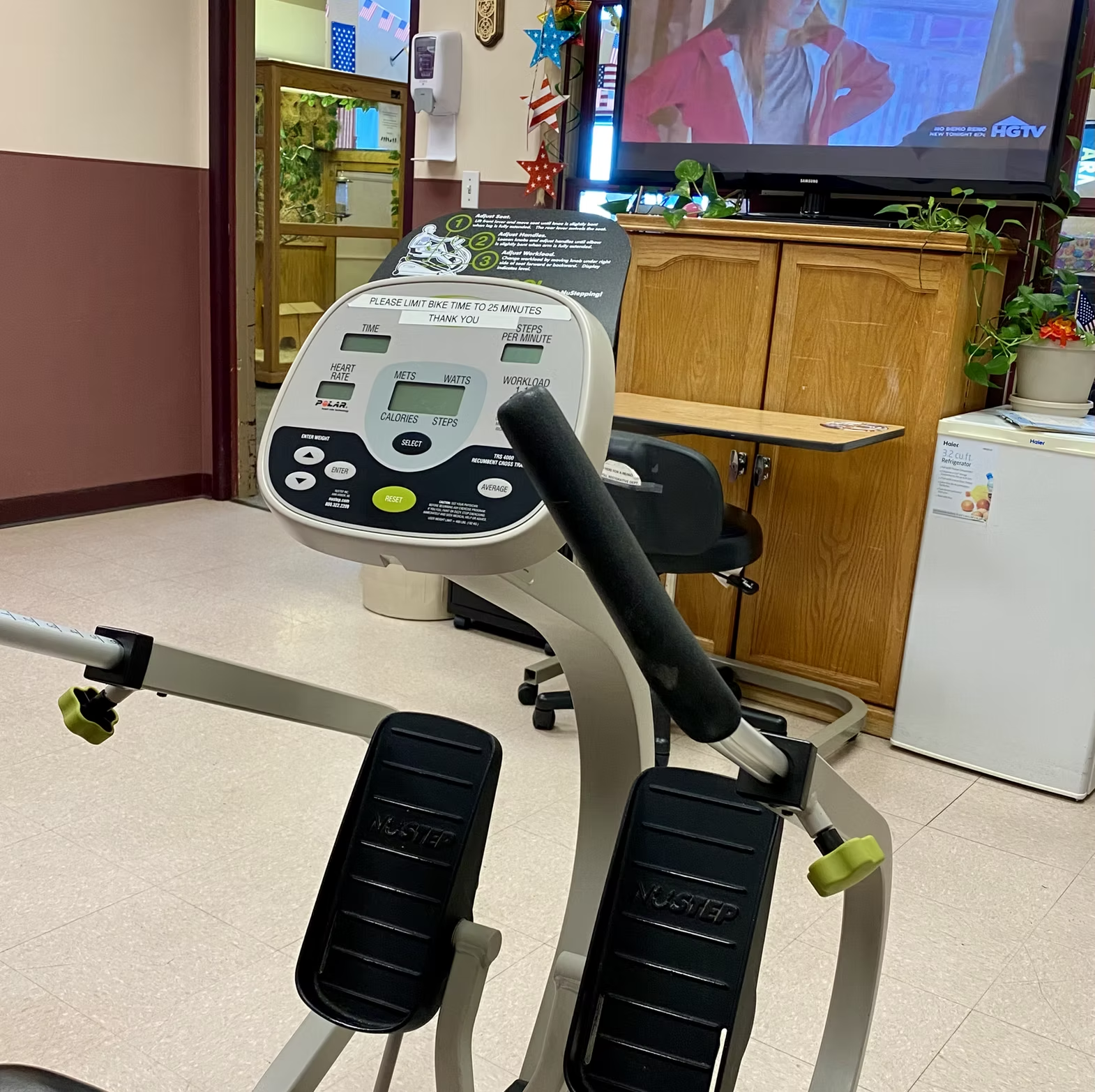 Exercise bike in a room with a TV, wooden cabinets, and plants. A door to another room is visible.
