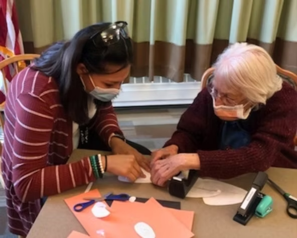 Two people wearing masks are working together on a craft project at a table with paper, scissors, and staplers.