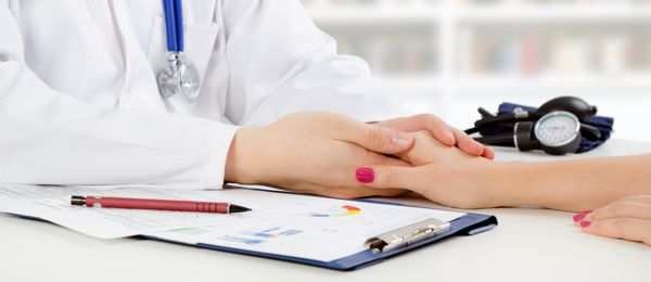 A doctor holding a patient's hand over a clipboard with papers. A stethoscope and blood pressure cuff are visible in the background.