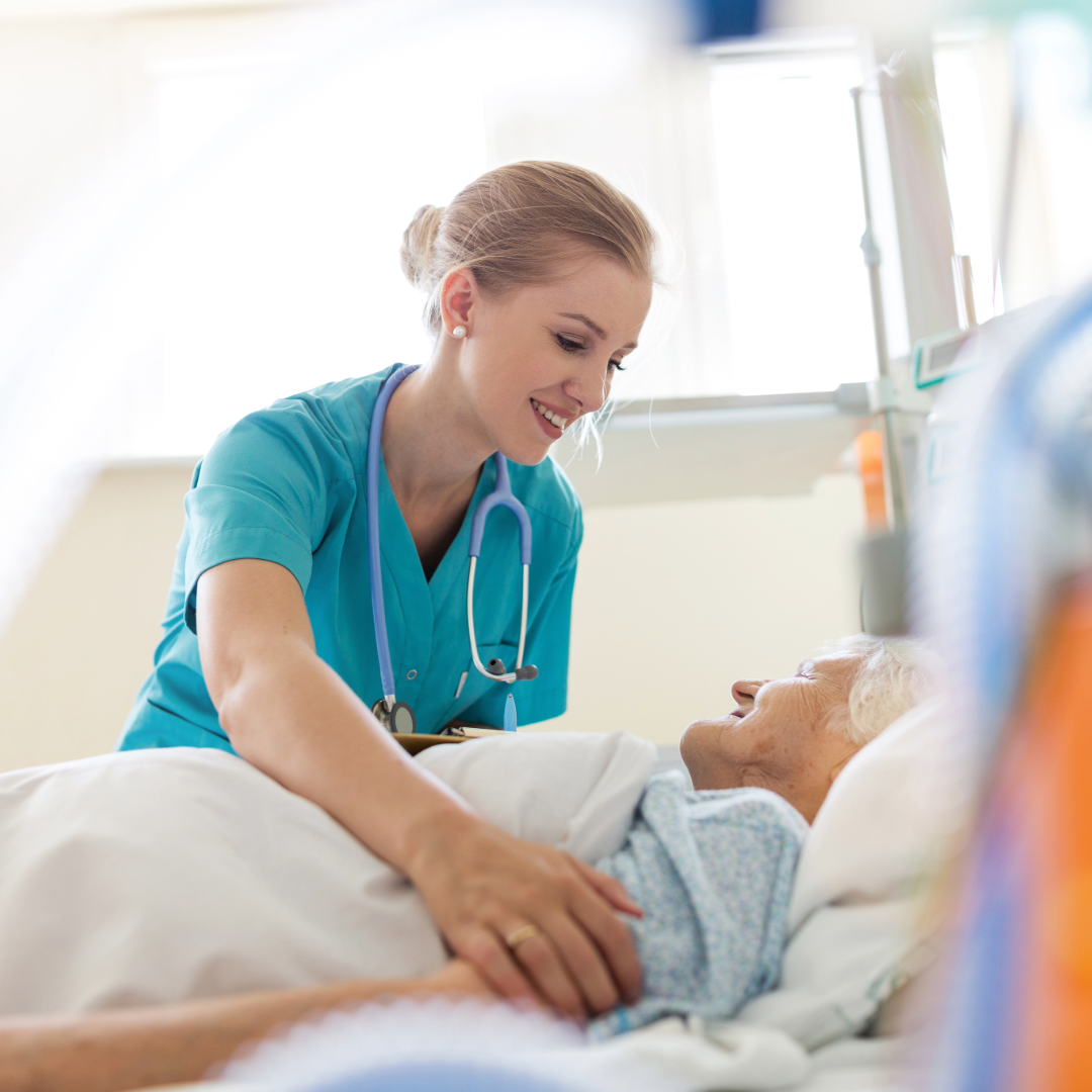 A nurse with a stethoscope smiles and cares for an elderly patient in bed at a hospital or care facility.