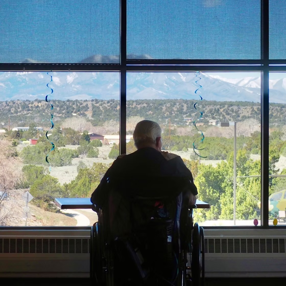 A person in a wheelchair looks out a large window at a mountainous landscape, with blue spirals hanging by the window.