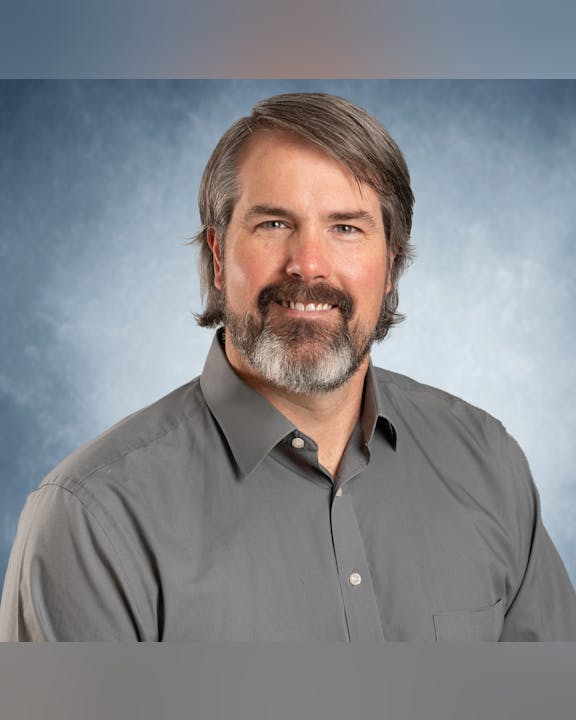 A man with a beard smiles in a portrait against a blue background, wearing a gray shirt.