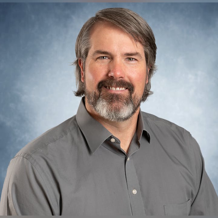 A man with a beard smiles in a portrait against a blue background, wearing a gray shirt.