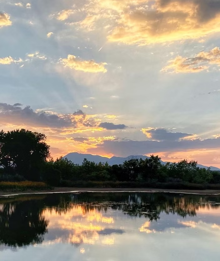 Sunset over mountains with trees reflected in a calm lake, surrounded by colorful clouds.