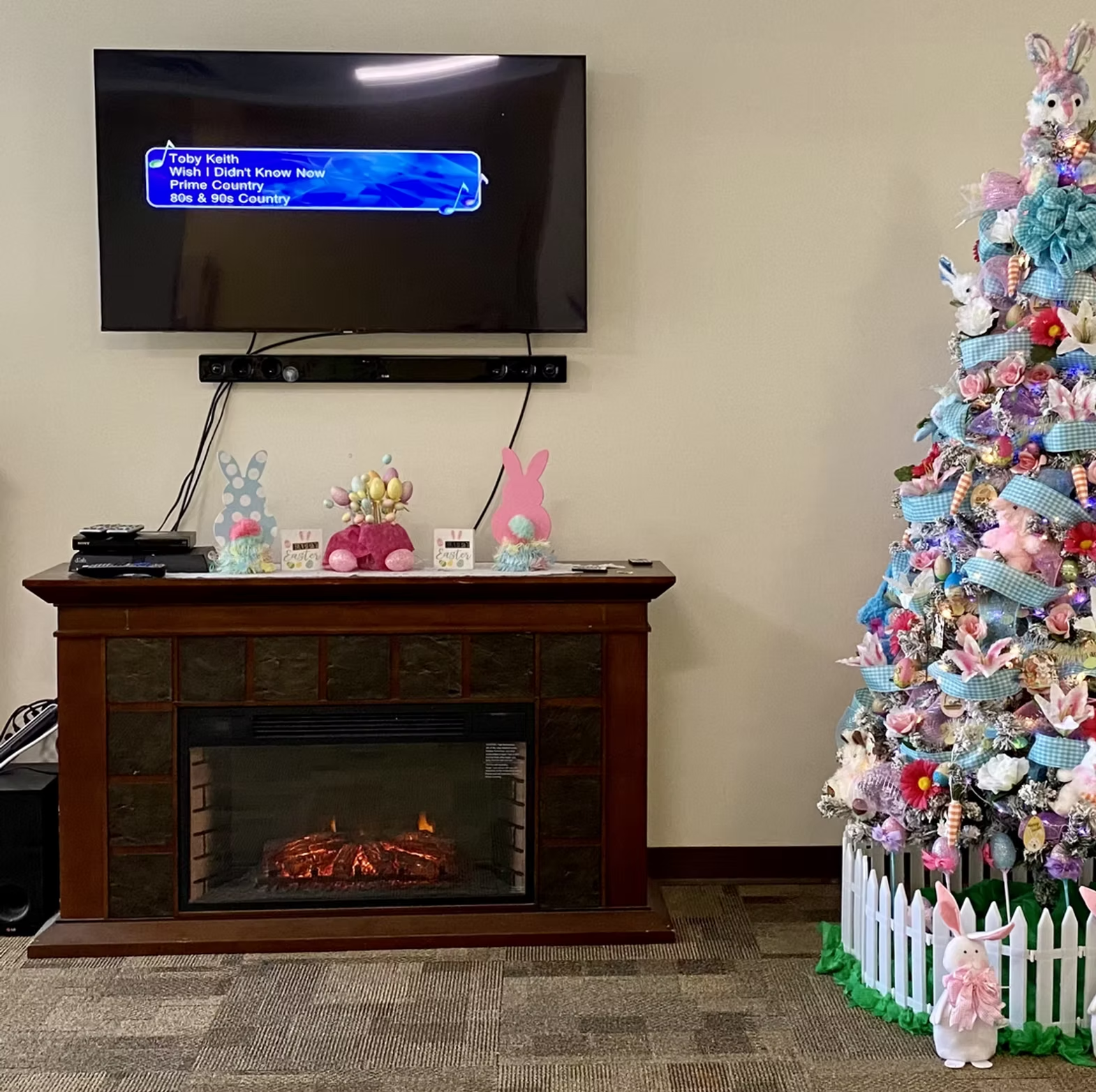 A TV above a fireplace, Easter-themed decorations including a tree with bunnies and eggs, and a song playing on the TV.