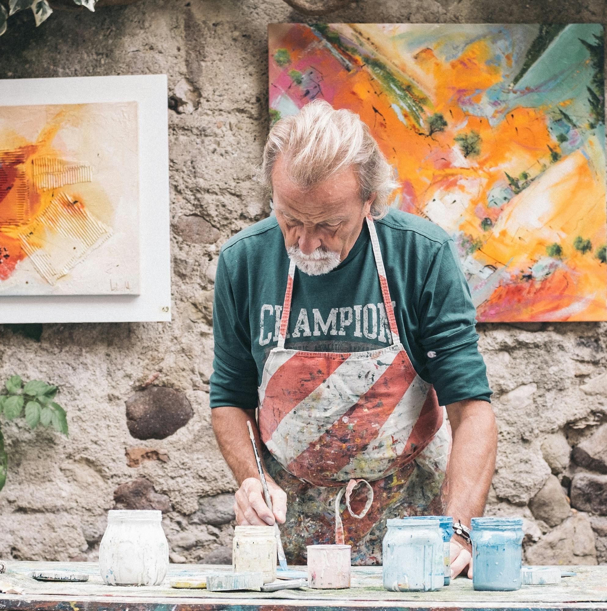 An artist painting outdoors, with colorful artworks and plants in the background.