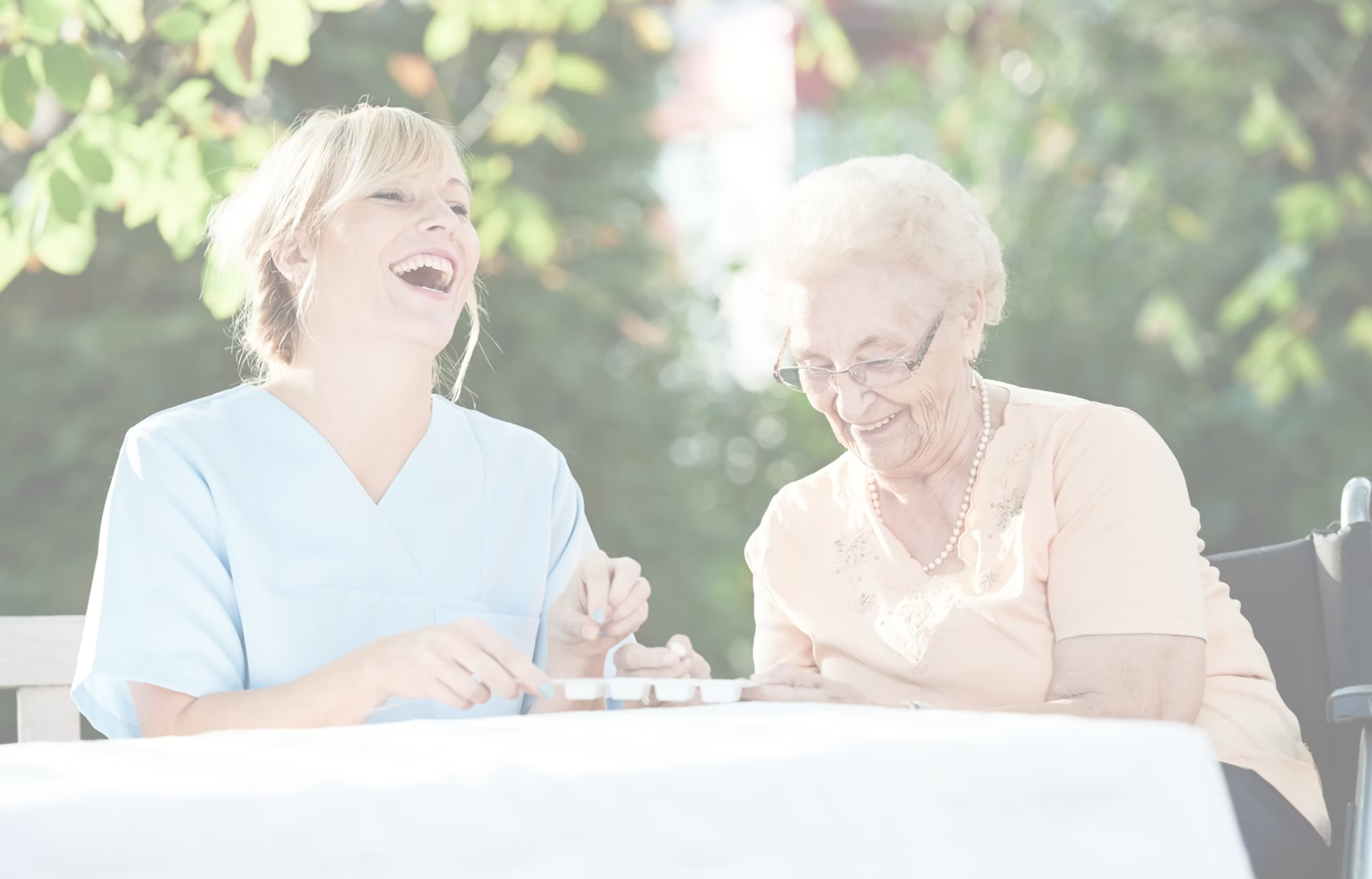 A caregiver and an elderly woman are sitting outside, laughing and enjoying each other's company.