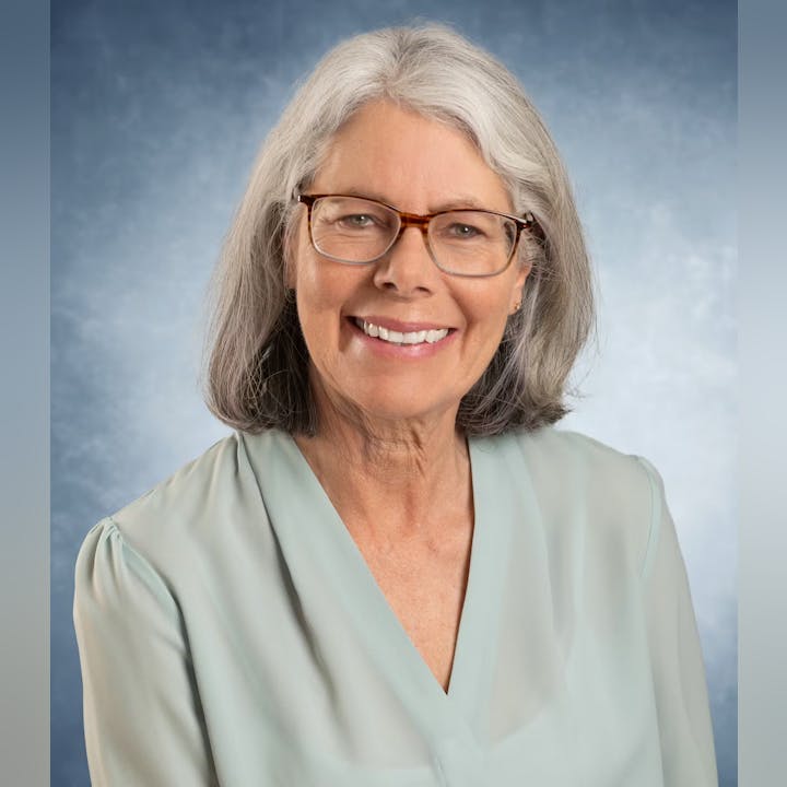 A smiling older woman with glasses and gray hair, wearing a light-colored blouse, against a blue background.