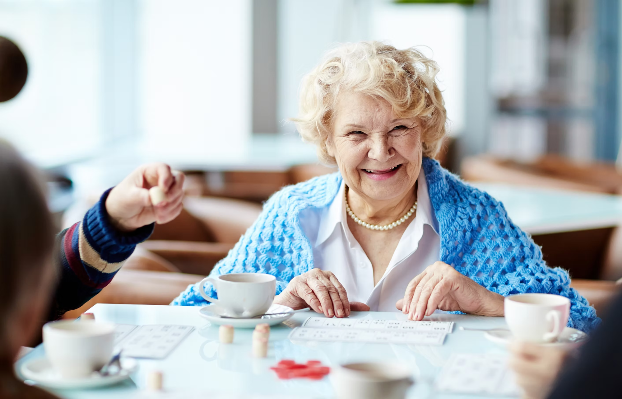 Smiling elderly woman playing a board game with friends, seated at a table with cups of coffee.