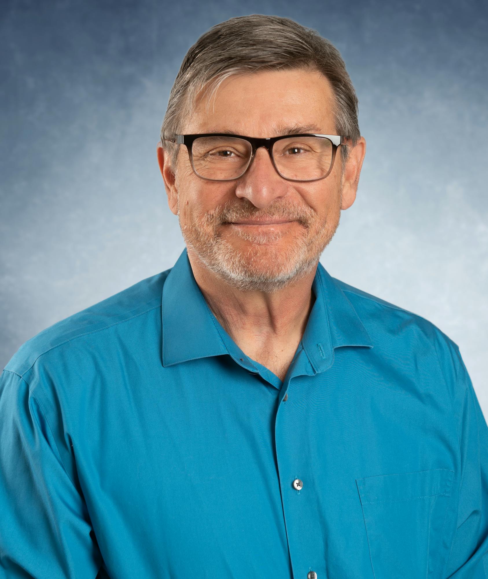 A smiling man with glasses, wearing a blue shirt, against a blue-gray background.