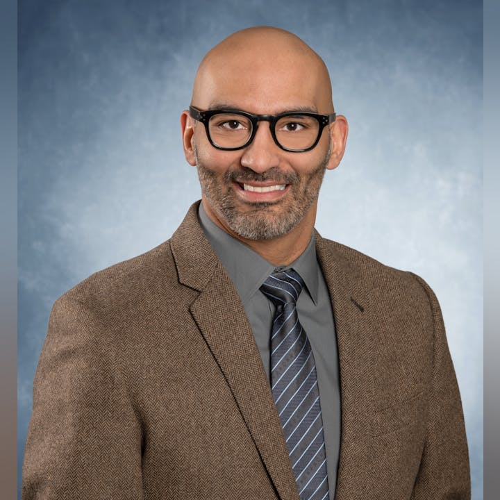 A person in a suit and tie, wearing glasses, smiling against a blue gradient background.