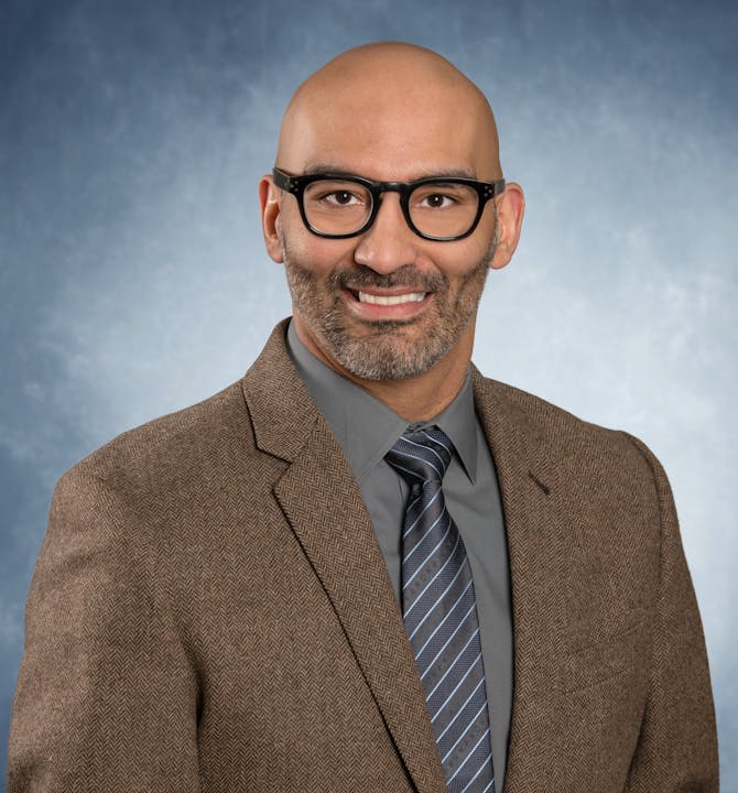 A person in a suit and tie, wearing glasses, smiling against a blue gradient background.