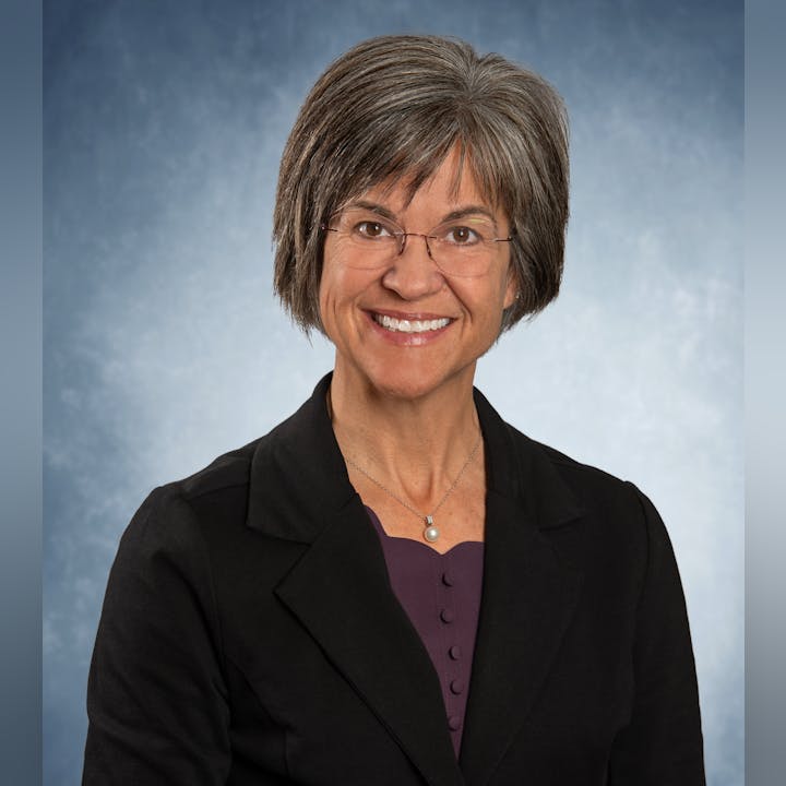 A person with short hair and glasses, wearing a black blazer over a purple top, smiles against a blue background.