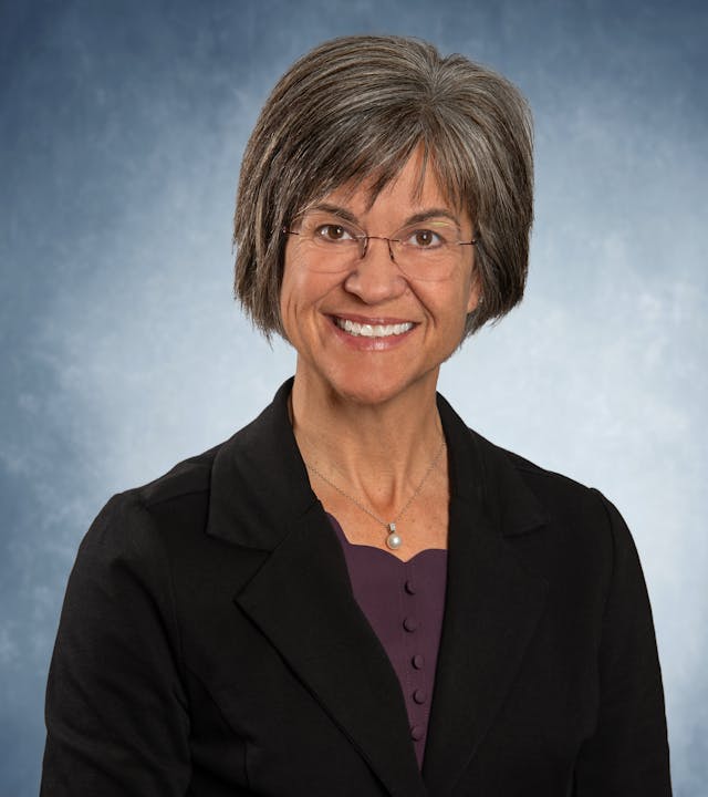 A person with short hair and glasses, wearing a black blazer over a purple top, smiles against a blue background.