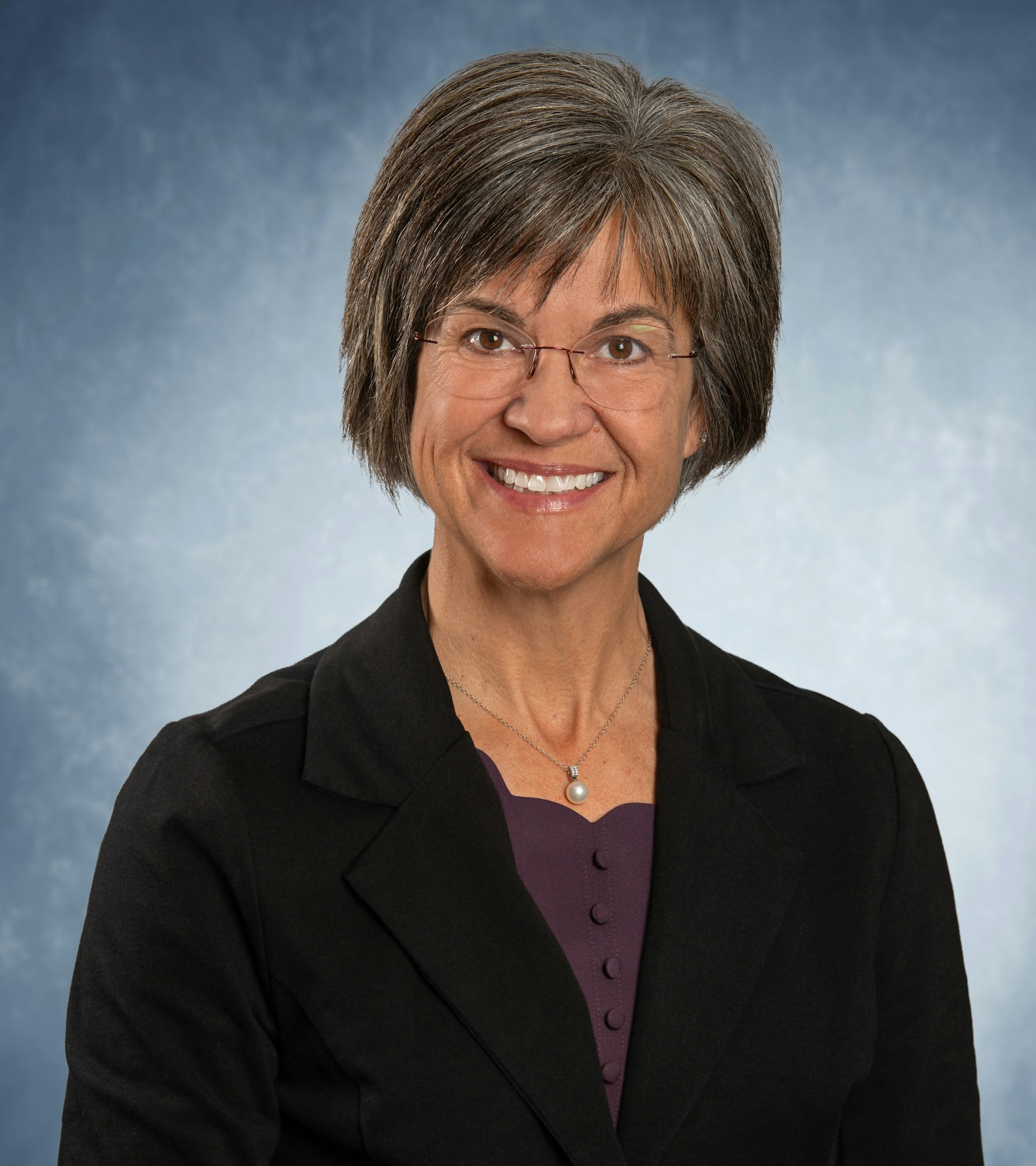 A person with short hair and glasses, wearing a black blazer over a purple top, smiles against a blue background.