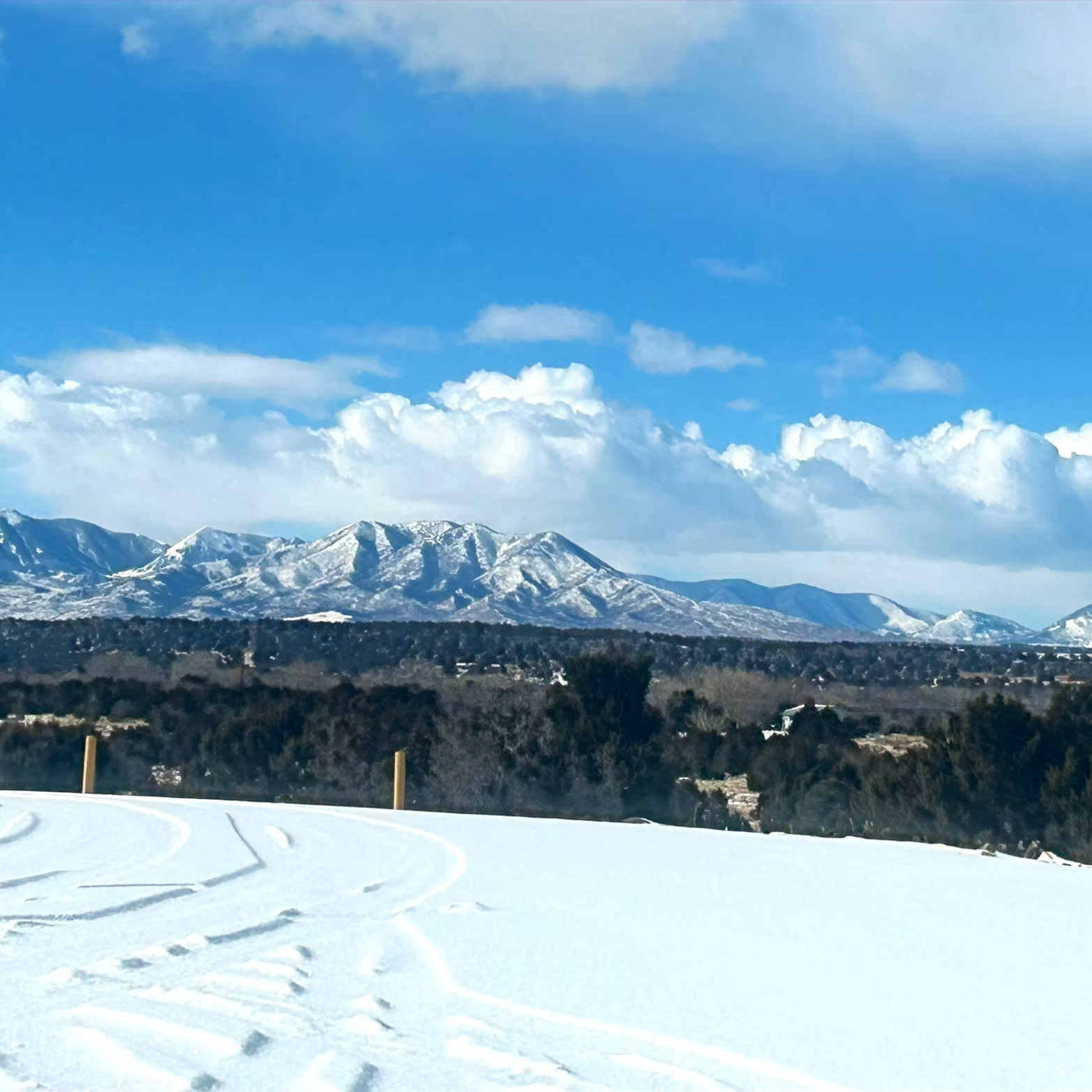 Snow-covered field with tire tracks, distant mountains under a bright blue sky and scattered clouds.