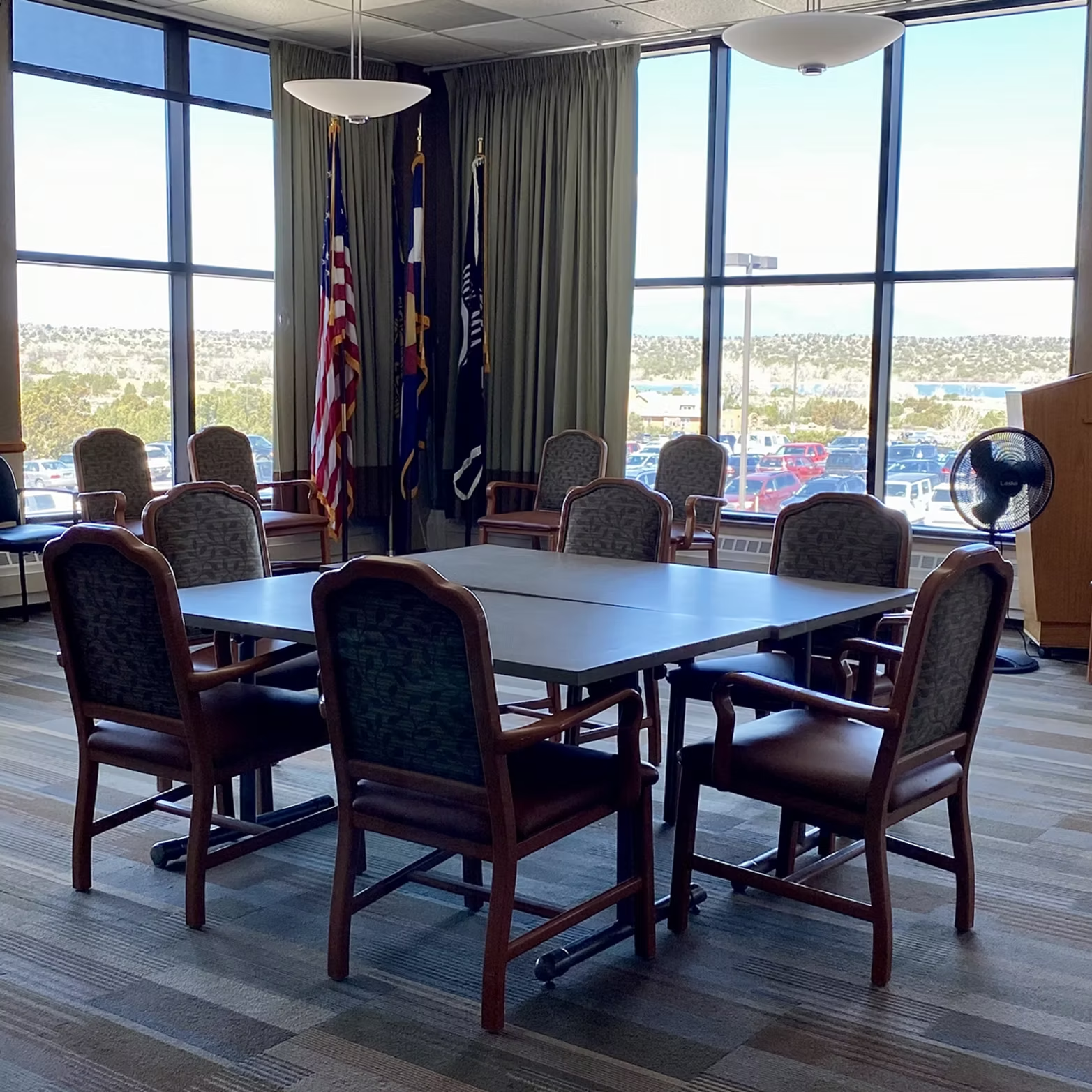 A conference room with a table, chairs, flags, large windows, a podium, and a fan.