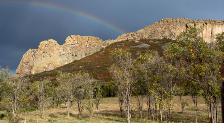 Rocky hill with a rainbow arching over, trees in the foreground, under a cloudy sky.