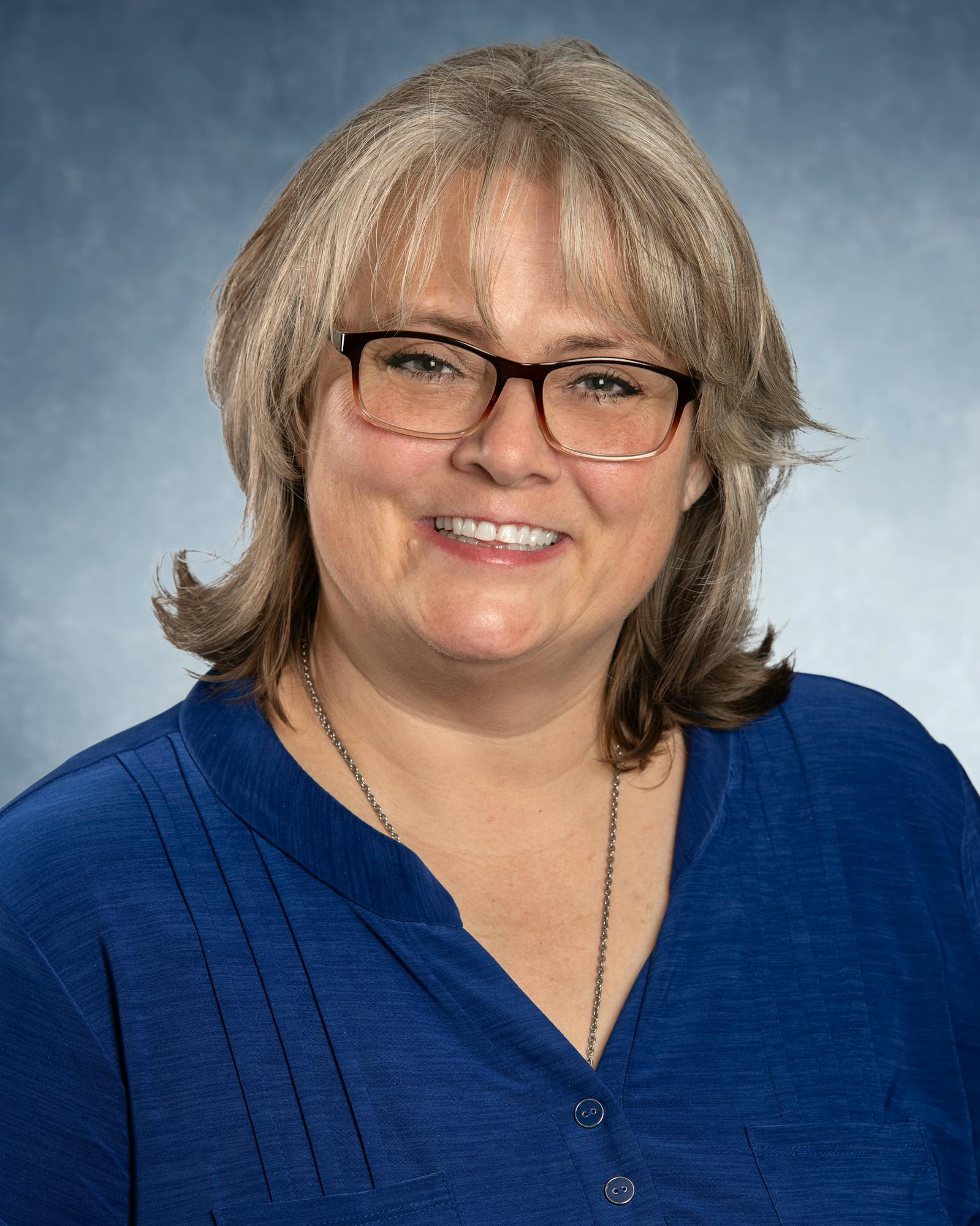 A person with glasses and gray hair smiles against a gray-blue background, wearing a blue shirt.