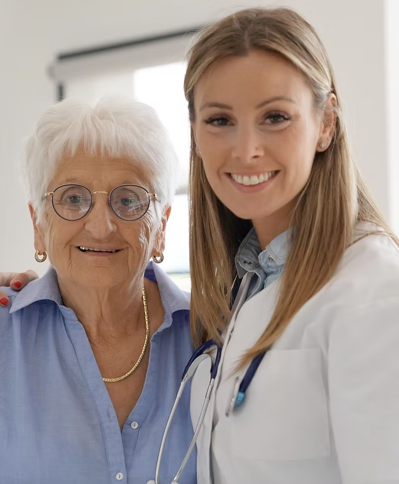 An older woman and a healthcare professional smiling together, the professional wearing a white coat and stethoscope.