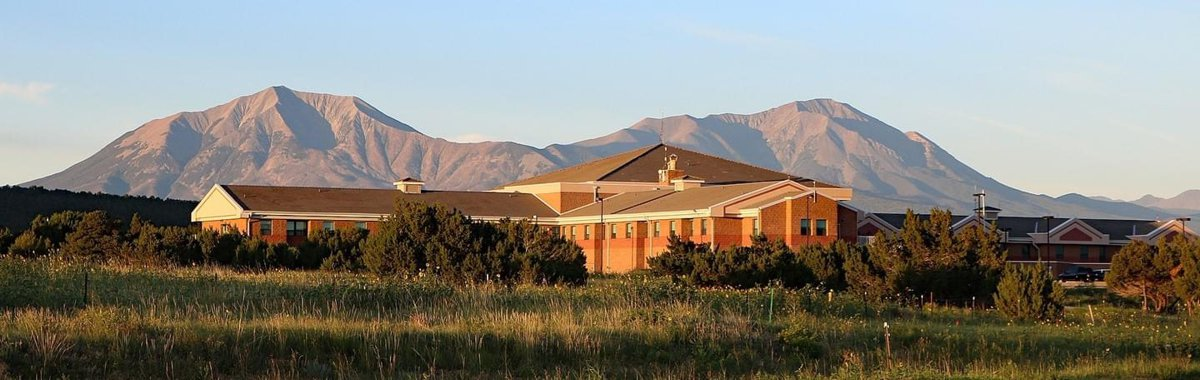 A large building with a mountain backdrop, surrounded by grass and trees under a clear sky.