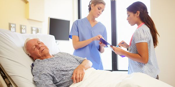 A patient lying in a hospital bed while two healthcare professionals discuss and take notes nearby.