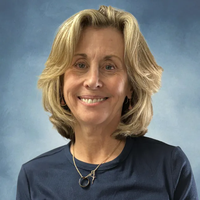 A smiling person with blonde hair, wearing a navy blue shirt and a necklace, is seated against a soft blue backdrop.