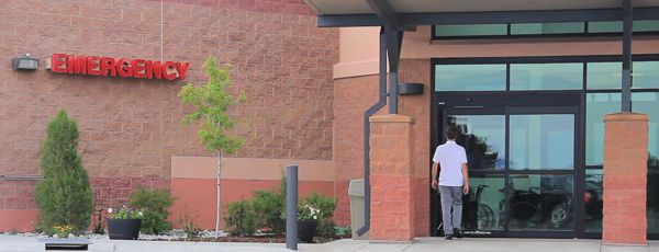 A person entering an emergency room entrance, with red "EMERGENCY" sign and brick walls in the background.