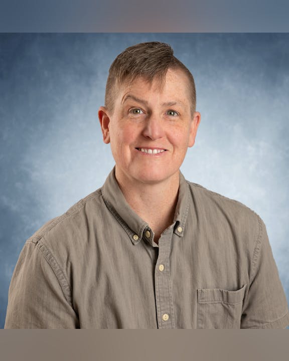 Person with short hair wearing a button-up shirt, smiling against a blue background.