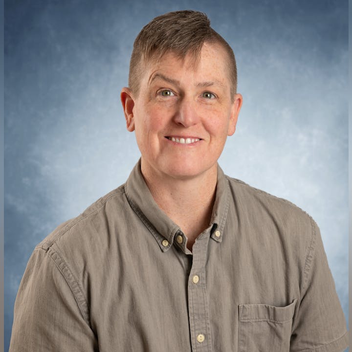 Person with short hair wearing a button-up shirt, smiling against a blue background.