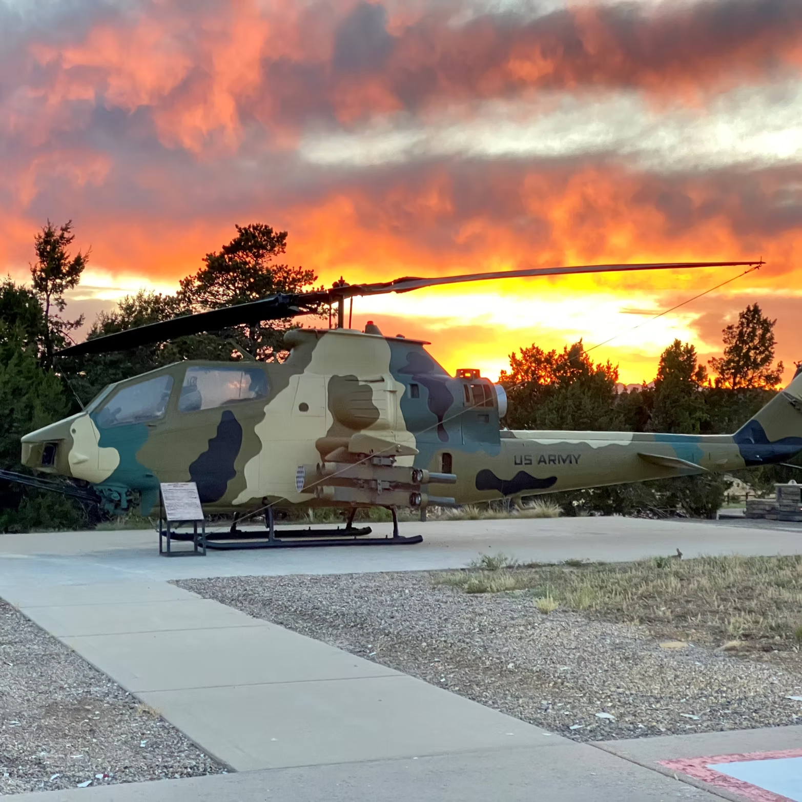 A camouflaged US Army attack helicopter on display at sunset with a fiery sky and trees in the background.