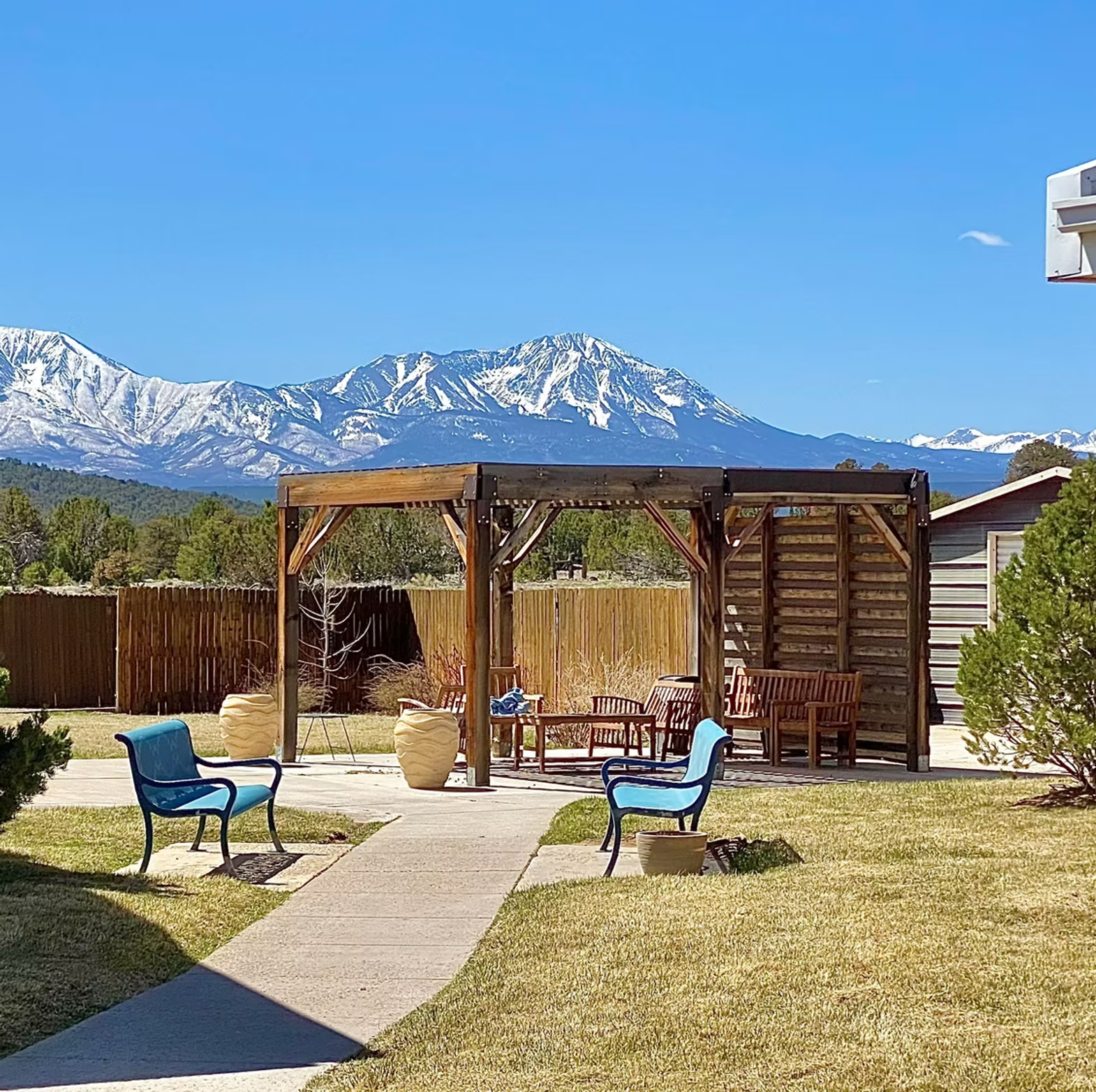 Outdoor patio with pergola, two blue chairs, snow-capped mountains in the background, clear blue sky.