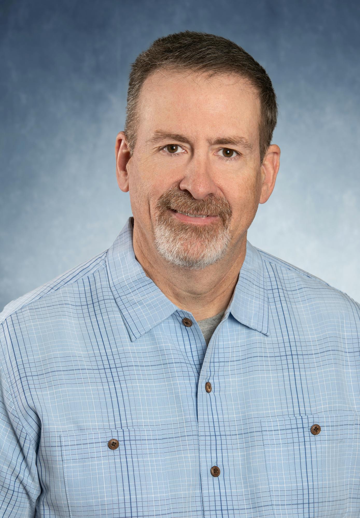 A person in a light blue plaid shirt with a neutral expression against a soft blue background.